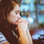 Woman praying with folded hands, reflecting on the All Knowing God who knows our thoughts, needs, and future.
