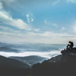 Person sitting high on a mountain overlooking the horizon, reflecting on Jesus as the Impregnable Rock and eternal refuge above life’s struggles.