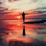 A silhouette of a person standing on a beach with arms raised in praise to God, set against the backdrop of a vibrant sunrise.