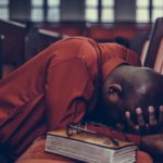 Man kneeling in prayer beside a Bible representing trust in Jesus, the Encouraging God, during times of hardship and discouragement.