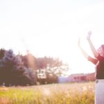 A woman standing in a sunlit field with her hands raised in praise, reflecting on God Who Contends For His People and rejoicing in His faithful protection.