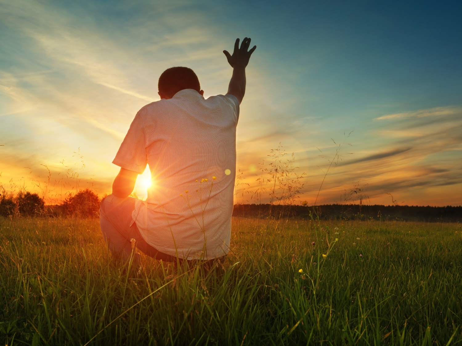 A man kneeling in a field praying with his hand raised