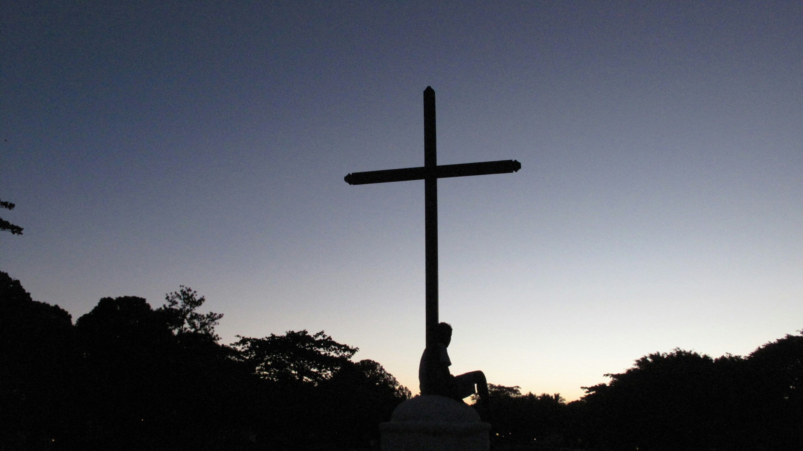 The silhouette of a person sitting by a cross.