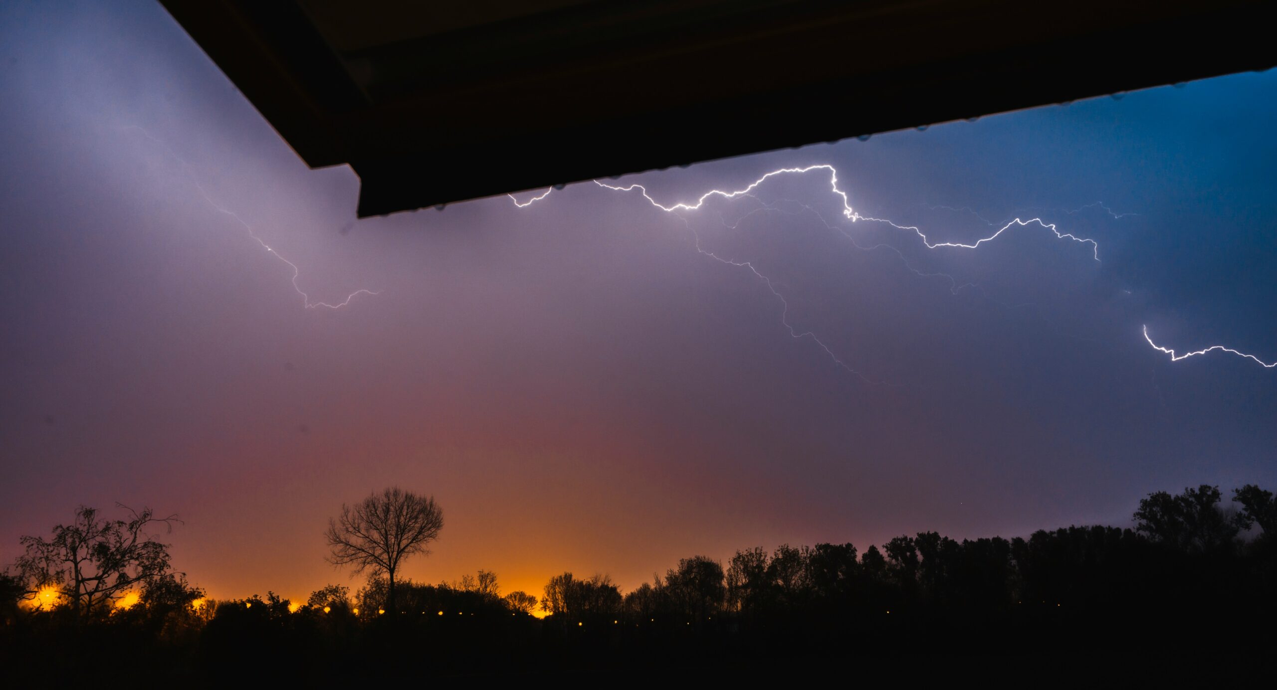 Part of a roof in the foreground and lightning/a storm in the background.