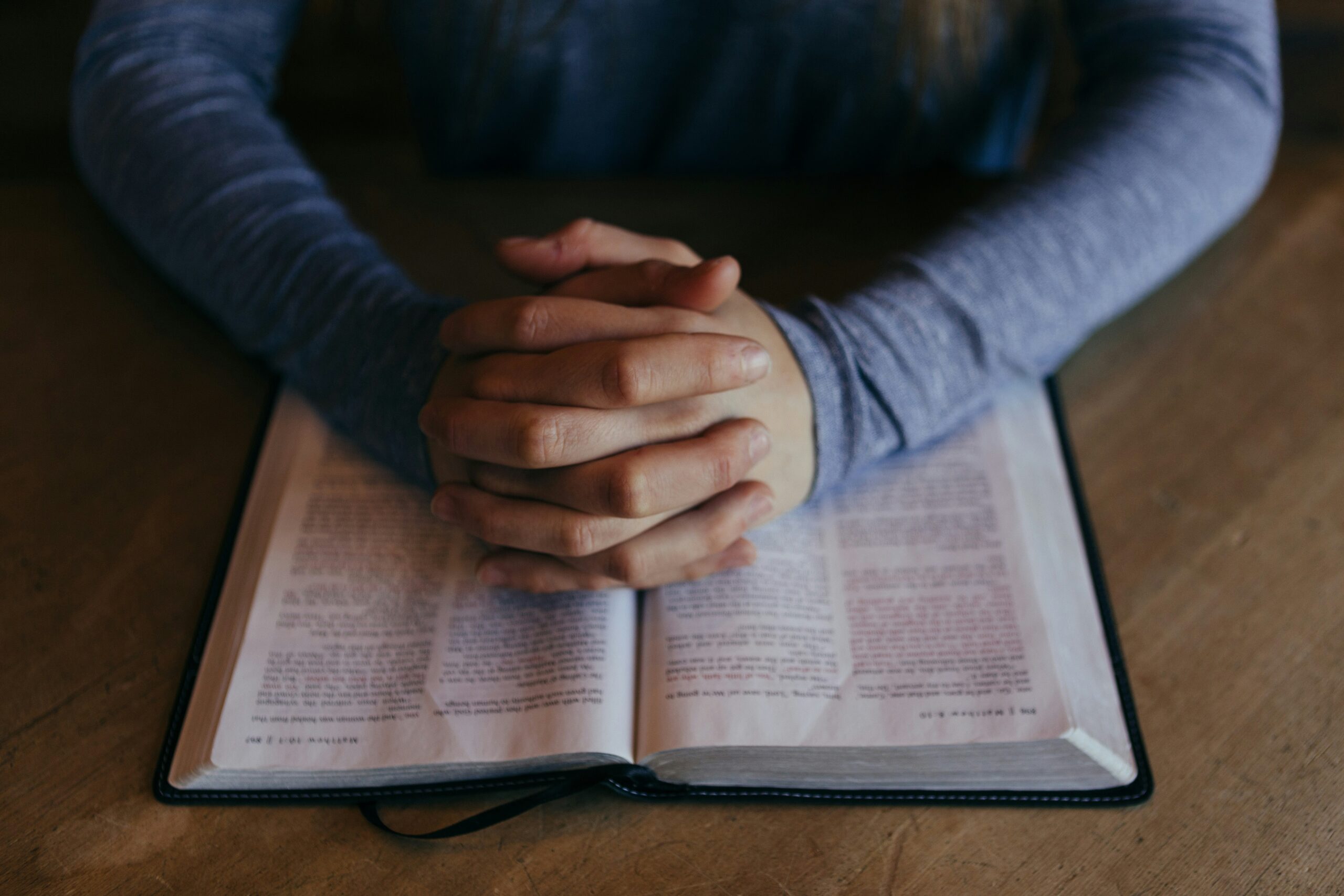 A man's hands with fingers interlaced in prayer over an open Bible.