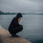 Person sitting alone by the water in a moment of discouragement, reflecting the need to trust the God of Hope who fills us with joy and peace.