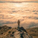 Person standing on a mountain top above the clouds, reflecting on the Majestic Glory of God and the hope of seeing Christ’s glory.