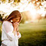 Woman praying in a sunlit field, reflecting on God’s Magnificent Promises and His divine nature through Christ.