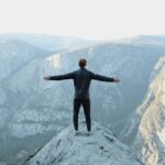 Man standing with arms outstretched on a mountain peak, reflecting on Jesus, the overcomer, who gives strength and peace in life’s challenges.